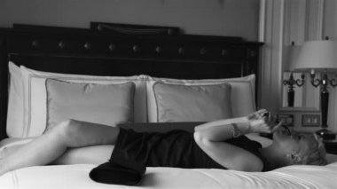 Lüks otel yatak odasında sıska kısa elbise takı güneş gözlüğü giyen güzel seksi sarışın iş kadını içki şarap toplantı dan önce dinlenmek moda trendi kırmızı ruj yastık çarşafları makyaj yapmak