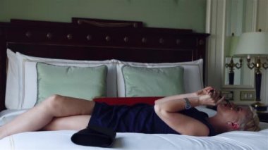 Lüks otel yatak odasında sıska kısa elbise takı güneş gözlüğü giyen güzel seksi sarışın iş kadını içki şarap toplantı dan önce dinlenmek moda trendi kırmızı ruj yastık çarşafları makyaj yapmak