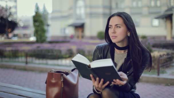 Mulher morena jovem bonita ler um livro no parque vento soprou seu cabelo, ao ar livre na primavera ou outono, tempo quente, uma menina vestida com uma jaqueta escura, de pé ao lado Bag — Vídeo de Stock