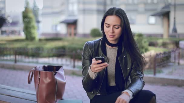 Krásná žena s tmavými vlasy, která sedí v parku a dívá se na telefon, čeká na schůzku nebo schůzku, napsala zprávu na ulici je dobré teplé počasí, jaro nebo pád, nosí černou bundu — Stock video