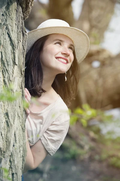 Ung kvinna med hatt titta runt ett träd — Stockfoto