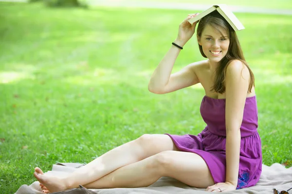 Mladá žena sedí v parku s knihou na hlavě — Stock fotografie