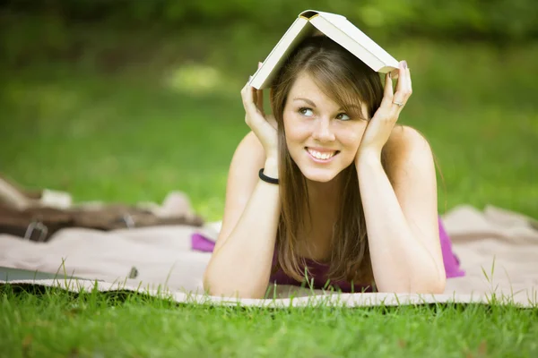 Mujer joven acostada en el parque sosteniendo el libro en su cabeza — Foto de Stock