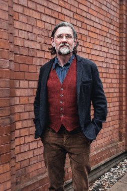 50 'li yaşlarda yakışıklı bir adam tuğla duvarın önünde duruyor.