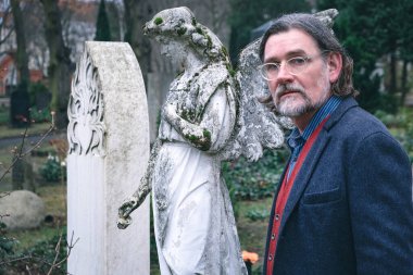 50 'li yaşlarda yakışıklı bir adam mezarlıkta bir meleğin yanında duruyor.