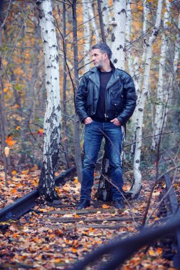Deri ceketli ve kotlu yakışıklı adam ormanda dikiliyor.