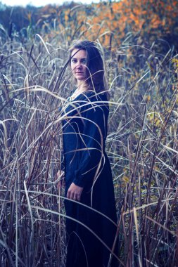 Siyah elbiseli esmer kadın sazlığın arasında duruyor ve üzgün görünüyor.