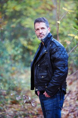 Deri ceketli ve kotlu yakışıklı adam ormanda dikiliyor.