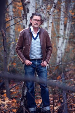 50 'li yaşlarda kahverengi deri ceketli yakışıklı bir adam ormanda dikiliyor.
