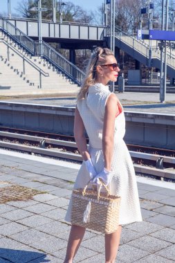 40 'larında beyaz elbiseli sarışın bir kadın tren istasyonunda duruyor.