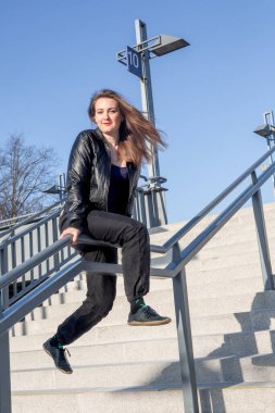 Deri ceketli genç bir kadın dışarıda merdivenlerde oturuyor.