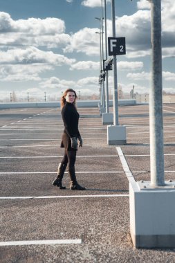 Siyah elbiseli ve botlu genç esmer kadın boş otoparkta duruyor.