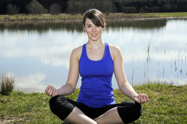 Young woman sitting by a lake in a lotus position — Stock Photo, Image