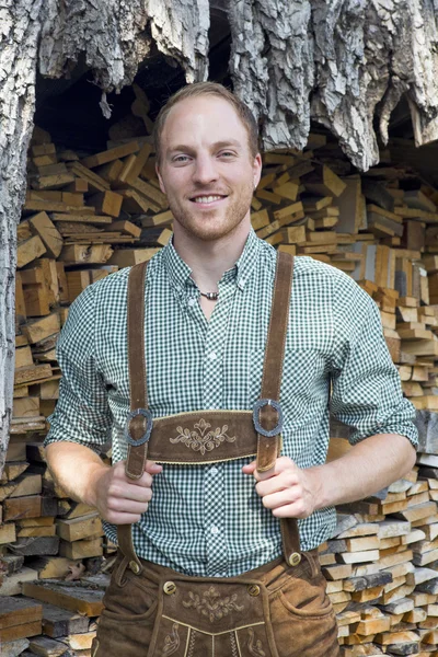 Junger Mann in bayerischen Lederhosen vor Brennholz — Stockfoto