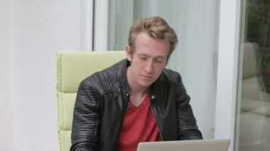 Genç sarışın adam dışında balkonda oturan ve büyük bir gülümseme ile laptop yazarak