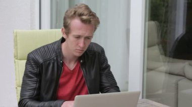 Genç sarışın adam dışında balkonda oturan ve dizüstü bilgisayarda yazarak