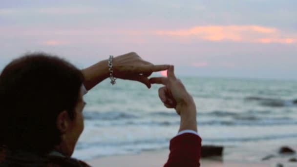 Женщина обрамляет солнце руками на закате на пляже — стоковое видео