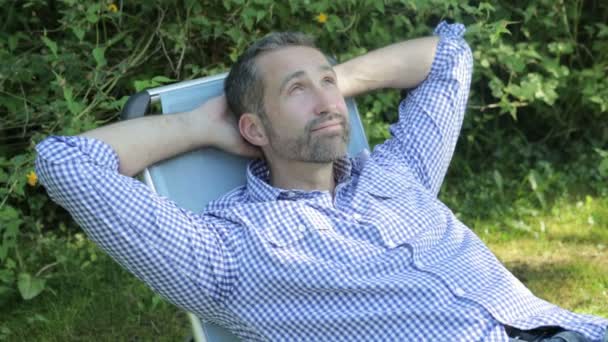 Hombre guapo sentado en una silla en un jardín y se relaja — Vídeos de Stock