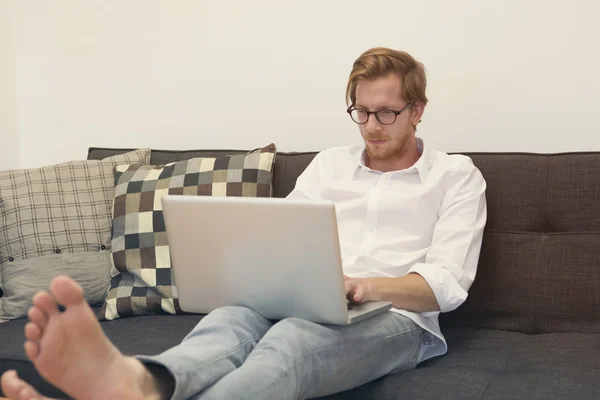 Ung man sitter barfota soffan med laptop — Stockfoto