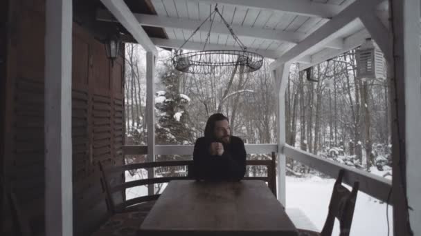 Όμορφος νεαρός άνδρας κάθεται στη βεράντα του ένα παλιό ξύλινο σπίτι κατά τη διάρκεια της χειμερινής περιόδου. — Αρχείο Βίντεο