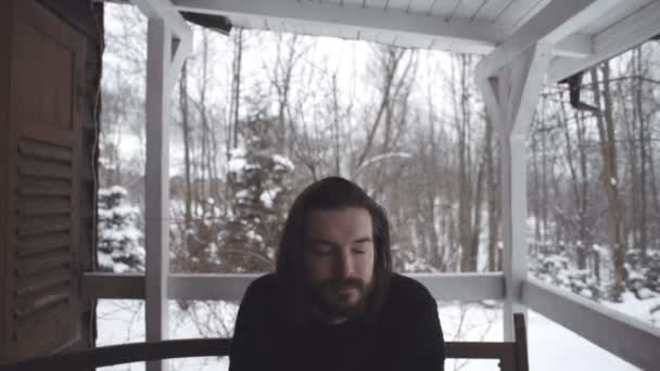 Knappe jonge man zit op de veranda van een oude houten huis tijdens winterseizoen. — Stockvideo