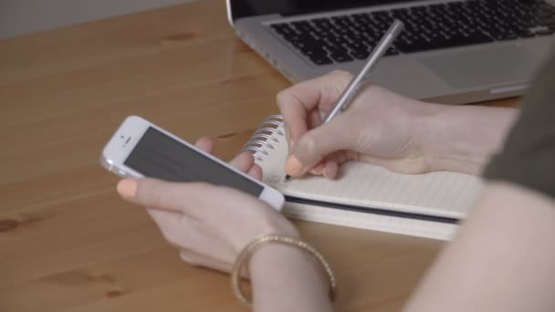 Mujer escribiendo a mano en la agenda y comprobando en un teléfono móvil en un escritorio . — Vídeos de Stock
