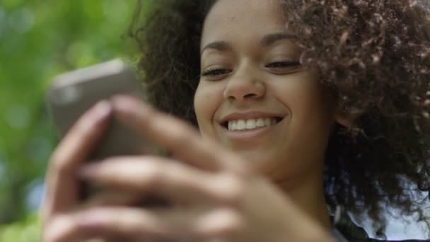 Belle jeune fille aux cheveux bouclés foncés en utilisant son téléphone portable, en plein air . — Video