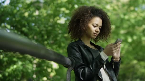 Menina bonita com cabelo encaracolado escuro usando seu telefone celular, ao ar livre . — Vídeo de Stock