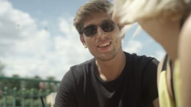 Genç neşeli grup arkadaşıyla sahilde gülüyor, söz ve ahşap koltuğa otururken soğuk içecekler içme - birlikte eğleniyor. 