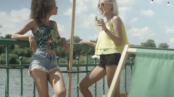 Schöne junge Mischlingsmädchen, die in der Nähe eines Sees stehen, reden, trinken und Spaß haben. — Stockvideo