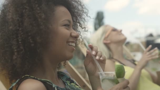Jóvenes hermosas chicas de raza mixta sentadas en tumbonas bajo el paraguas y disfrutando de vacaciones . — Vídeo de stock