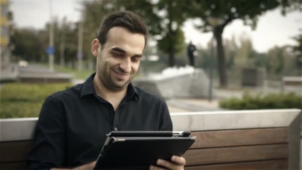 Homem atraente feliz usando tablet PC fora em um banco de parque — Vídeo de Stock