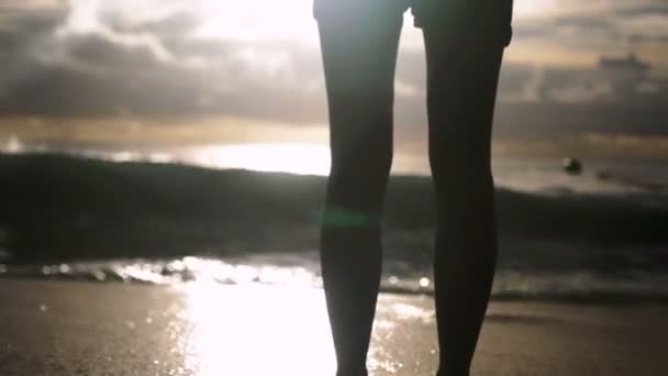 Jovem mulher de shorts de pé sozinha na praia ao pôr do sol enquanto a água lava os pés . — Vídeo de Stock