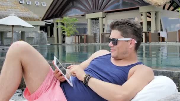 Man wearing sunglasses sitting by pool with digital tablet in tropical resort. — Stock Video