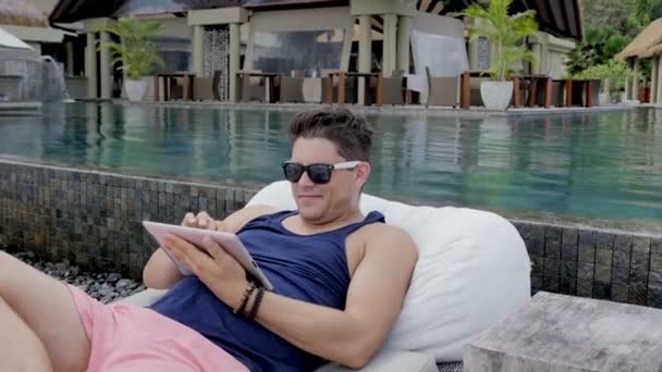 Hombre joven casual sentado junto a la piscina con tableta digital en el resort tropical . — Vídeo de stock