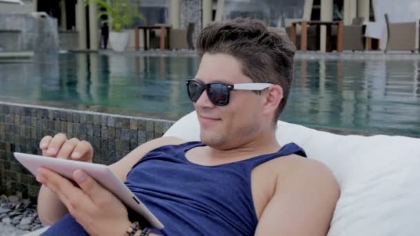 デジタル タブレットのトロピカル リゾートで、プールのそばに座ってカジュアルな若い男. — ストック動画