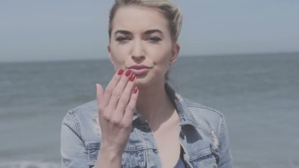 Heureuse femme blonde en veste en denim envoyant un baiser sur une plage solitaire . — Video