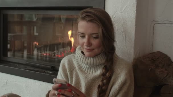 Hermosa mujer usando un teléfono celular en casa en el calor de una chimenea . — Vídeo de stock