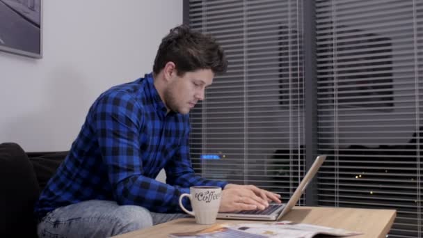 Молодий кавказький чоловік працює на столі з ноутбуком. Усміхнений молодий чоловік використовує ноутбук у вітальні . — стокове відео