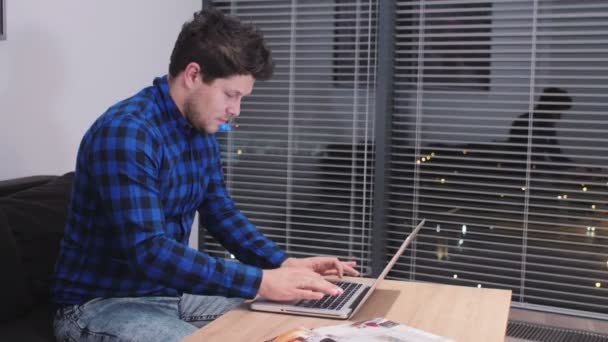 一个年轻的白种人男子在一张书桌和一台笔记本电脑上工作。使用笔记本电脑在客厅里的年轻男人微笑着. — 图库视频影像