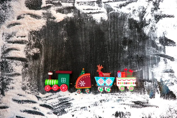 Eine Spielzeugeisenbahn aus Holz trägt Geschenke und weihnachtliche Utensilien auf dunklem, schneebedecktem Hintergrund. Image für ein dekoratives Weihnachtsfeiertagskonzept. Winterrahmen. Freier Raum zum Kopieren. — Stockfoto