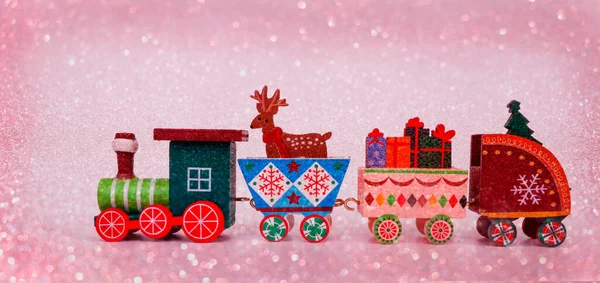 Eine Spielzeugeisenbahn aus Holz trägt die Attribute eines Winterurlaubs auf rosa Schneehintergrund. Postkarte für die Weihnachtsfeiertage. Image für ein dekoratives Weihnachtsfeiertagskonzept. — Stockfoto