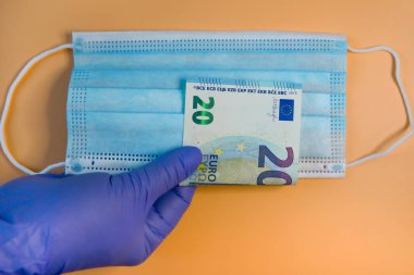 Eldivenli eller, tıbbi maske geçmişine karşı yirmi avroluk banknot tutuyor. Kıtlık kavramı ve bir salgın sırasında kişisel koruma ekipmanlarının maliyetinin artması..