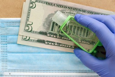 Eldivenli bir el, yüz maskesi ve beş dolarlık banknotlara karşı dezenfektan tutar. Koronavirüs salgını sırasında evi güvenli bir şekilde terk etmek için koruyucu ekipman kullanma maliyeti kavramı.