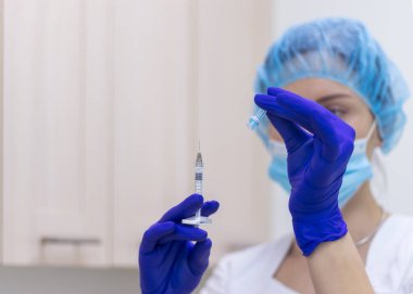 Beyaz üniformalı ve steril mavi eldivenli bir doktor şırıngayı iğneyle açıyor. Eller yakın çekim. Seçici odak