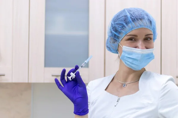 A female doctor wearing gloves and a mask holds a syringe with a needle. The concept of vaccination against dangerous diseases, including coronavirus. Selective focus, blurred background.