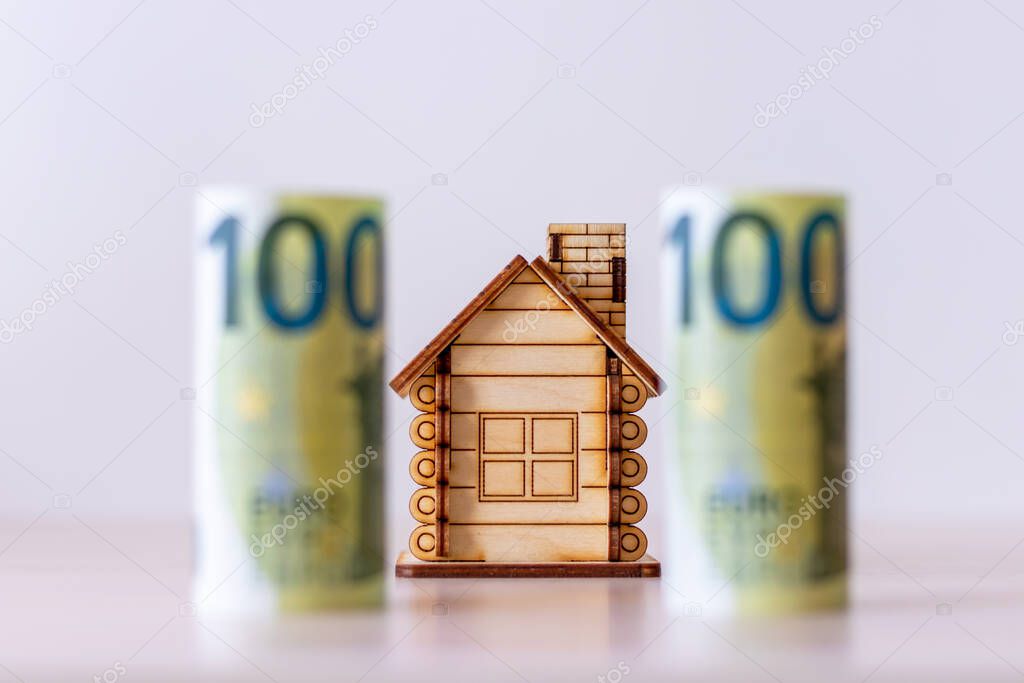 A wooden house on the background of blurred paper banknotes with a face value of 100 euros. The concept of the budget of the real estate market. Investment in construction. Mortgages and debts