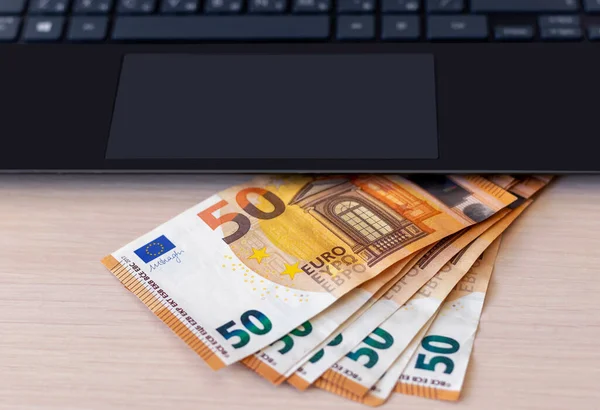 A black laptop and paper money worth 50 euros spread out in a fan on the table. The concept of online work and investment