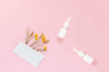 Tek kullanımlık koruyucu bir maskenin altında yabani çiçek ve otlar ve açık pembe bir arka planda burun spreyinin maketi. Mevsimsel alerjilerin korunması ve tedavisi kavramı