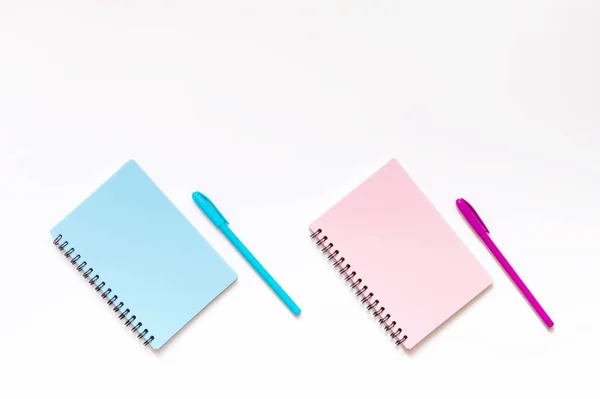 Beyaz arka planda iki defter ve pembe ve mavi renkli tükenmez kalemler. Yaratıcılık ve okul için ofis malzemeleri minimalist bir kavram. Düzen, üst görünüm, kopyalama için boş alan, düz uzanma